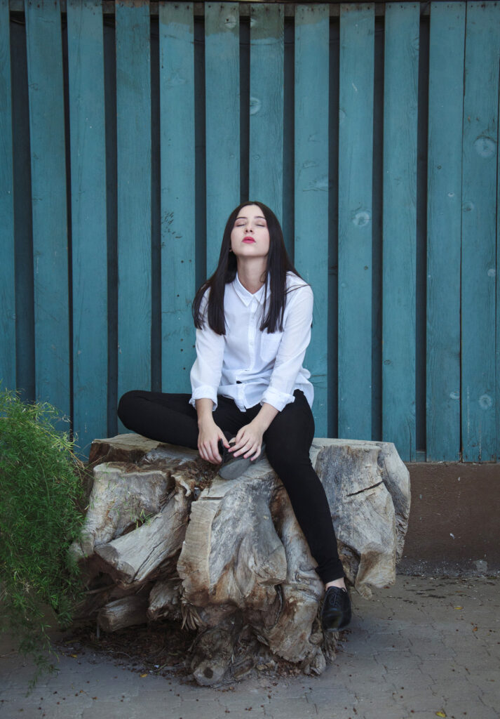 Gabriela Inzunza sits contemplatively on a weathered log against a teal wooden backdrop with a hint of greenery to her left.